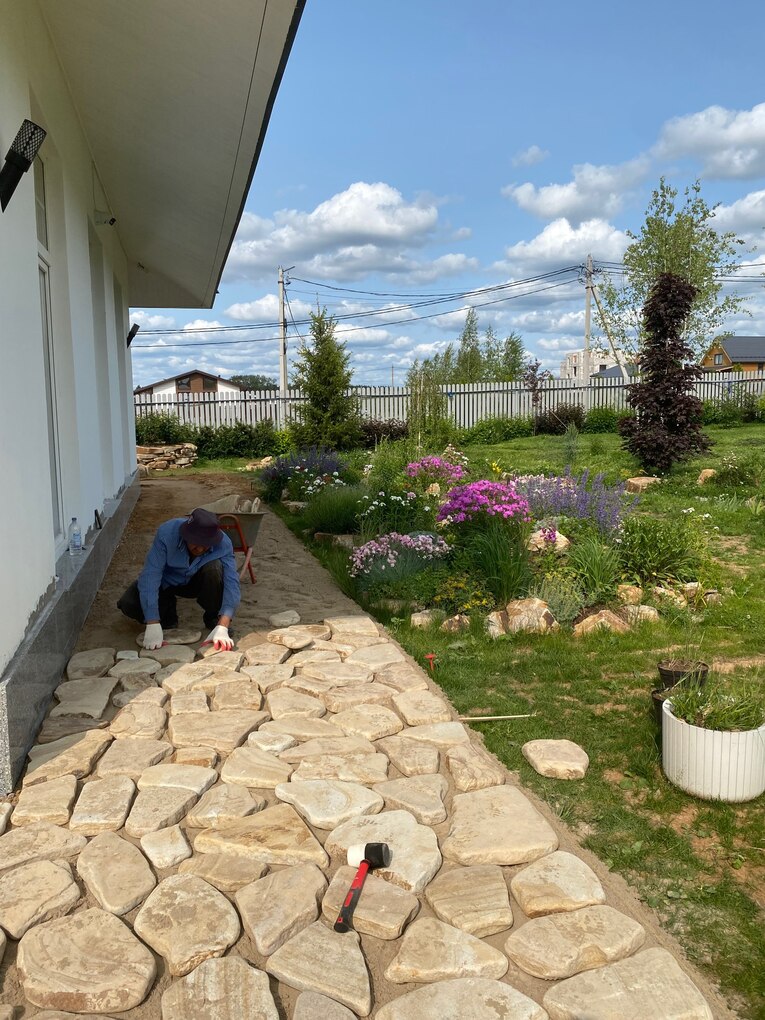 Рабочий выполняет укладку природного камня на дорожке рядом с цветником