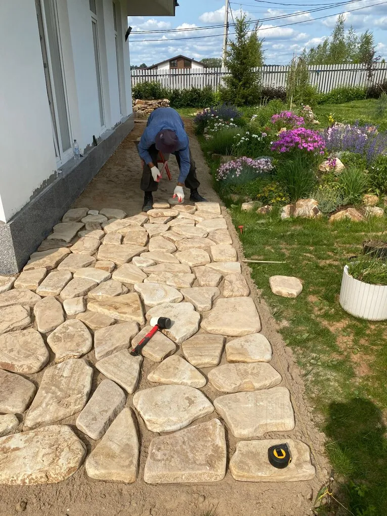 Мастер выравнивает плитняк на дорожке вдоль фасада дома